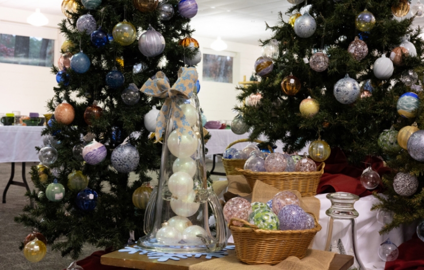 Image of two trees covered with ornaments with a small table in front. On top of the table is a clear glass cloche with ornaments stacked inside with a bow tied on top and two baskets filled with more ornaments to the right of it.