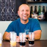 Carl Alfaro, co-owner of Conclave Brewery, smiling behind a selection of beers set up to taste.
